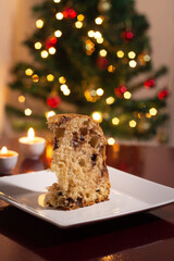 Fototapeta na wymiar Uma fatia de panetone dentro do prato, duas velas acesas e as luzes da árvore de natal ao fundo. Comida típica de Natal.