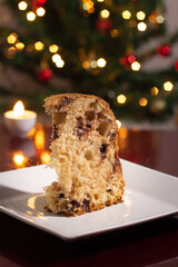 Uma fatia de panetone dentro do prato, duas velas acesas e as luzes da árvore de natal ao fundo. Comida típica de Natal.