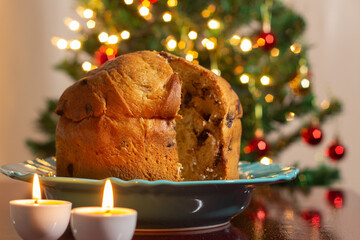 Fototapeta na wymiar Um panetone com duas velas acesas e as luzes da árvore de Natal borradas ao fundo. Comida típica de Natal.