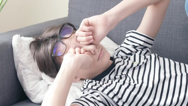 Tired teenager with glasses lying on sofa at home and rubbing his eyes. Tiredness, weakness after a hard day, resting at home, midday-napping, suffering from tired eyes, ocular diseases