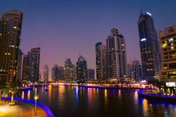 Fototapeta premium Dubai, UAE - 12th october, 2022: Dubai Marina skyline with illuminated sky scrappers,buildings and moving boats showing reflection on water captured from Marina Mall , Dubai, UAE.