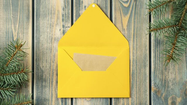 Stop motion video of bright yellow opening envelope on gray wooden background with branches of Christmas tree. Kraft paper card is taken out of new year envelope and opened. Copy space. Top view