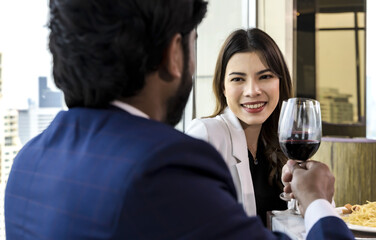 Smiling Asian woman drinking wine celebrate dating anniversary with indian boyfriend on rooftop restaurant, couples having surprise Dinner 