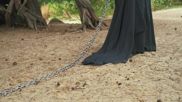 fantasy medieval goth woman holding pulling iron chains stretches across ground. Girl witch princess walks through dark forest. long black dress silk train. Back view. Concept slavery captive prisoner