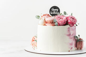 Birthday cake with white cream cheese frosting decorated with red roses and pink macaroons on the...