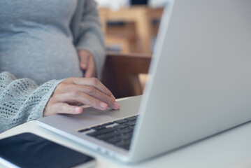 Fototapeta premium Pregnant woman working on laptop. Cropped image of pregnant businesswoman sitting at table typing on laptop at her working place in office