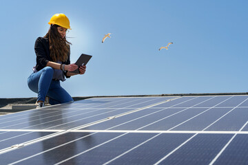 Ingegnere donna con caschetto protettivo giallo verifica le condizioni di un impianto fotovoltaico sul tetto di una casa con un tablet in mano - obrazy, fototapety, plakaty