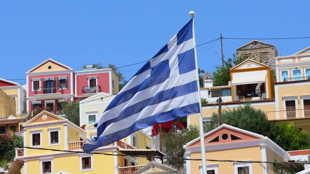 Greek flag flutters in the wind against the backdrop of the traditional houses of Symi island Greece