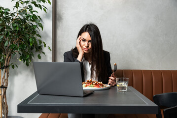Young frustrated business woman trying to eat her meal on lunch break in cafeteria but she cant because of constant interruptions by video call and e-mails from her work. Annoyed hungry female - obrazy, fototapety, plakaty