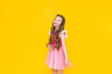 A little princess in a pink dress with curly hair is smiling broadly. A beautiful, charming child on a yellow isolated background.