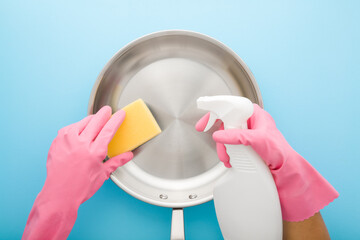 Woman hands in pink rubber protective gloves holding white detergent bottle, yellow sponge and washing stainless frying pan on light blue table background. Closeup. Pastel color. Top down view.