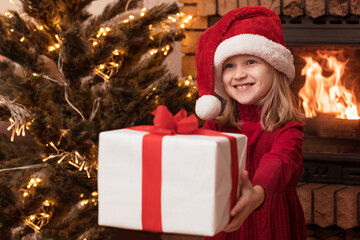 Fototapeta na wymiar Santa Girl Child Giving Christmas Present Gift Box in Christmas Home Atmosphere near Christmas Fireplace and Christmas tree