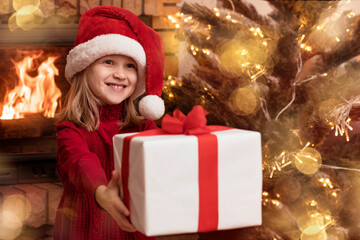 Fototapeta na wymiar Christmas Child in Santa Hat Give Christmas Gift Box in Cristmas Background with New Year Tree Lights and Fireplace. Copy Space