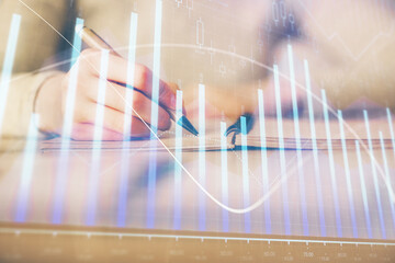 Hands writing information about stock market in notepad. Forex chart holograms in front. Concept of research. Multi exposure