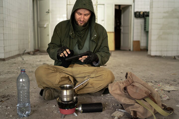 a dirty tired man sits on the floor in an abandoned room, warms her hands over a gas burner and prepares dinner for herself. Post-apocalypse. hiking in a post-apocalyptic world.