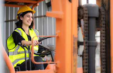 Female foreman wears hard hat driving forklift at shipping container yard, portrait. Multiracial industrial engineer woman drives reach stacker truck to lift cargo box at logistic terminal dock.