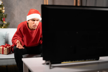 Young handsome man watching TV on couch at home. Arabic guy in Santa hat is argueing with TV. He is dissatisfied and waving with his hands. Christmas eve, home cinema