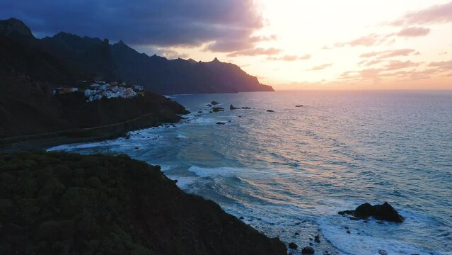 Deep dark night ocean water splash against mountain shore at sunset cloudy colorful sky. Little hillside village Almaciga. Amazing nature landscape. Tenerife north Canary island.