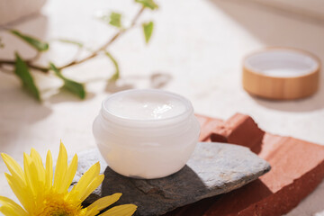 Obraz na płótnie Canvas White cream jar on natural stones podium and yellow flower on white background with light and shadows. Organic spa cosmetic beauty product. Closeup