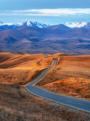 Vertical view of a picturesque road stretching into the distance through the picturesque autumn hills. Winding asphalted clean road stretches into the distance to the snow-capped mountains.