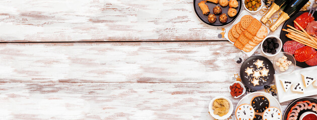 New Years Eve theme appetizer side border. Above view on a white wood banner background. Charcuterie board, champagne, clock cookies, tuxedo brie cheese and a variety of party food. Copy space.