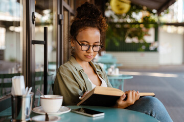 Hispanic woman in eyeglasses drinking coffee and reading book at cafe