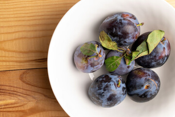 Several juicy black plums in a white plate, close-up, on a wooden table, top view.