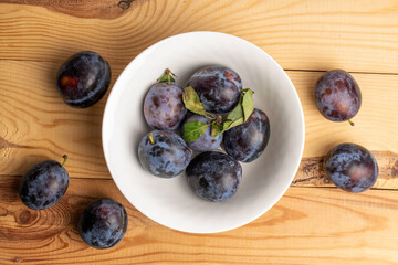 Several juicy black plums in a white plate, close-up, on a wooden table, top view.