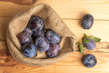 Several juicy black plums in a jute bag, close-up, on a wooden table, top view.