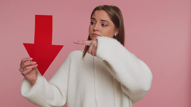 Upset young woman showing red arrow pointing down, concept of downgrade, unsuccessful business, fall of stock market money exchange rate, bankruptcy fail. Sad girl isolated on pink studio background