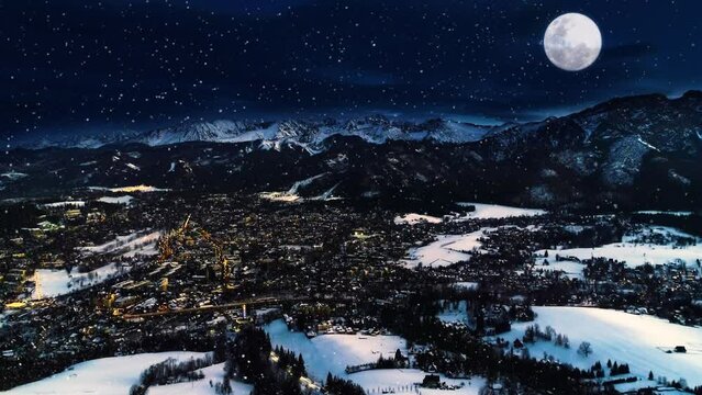 Snowy winter night, epic aerial view. Magical winter in the mountain village. Illuminated alpine city covered with snow. White mountains, ski resorts and big shining moon. Christmasy snowy landscape.