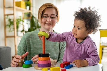 Mother looking at a child playing with an educational didactic toy. Young woman and child playing...