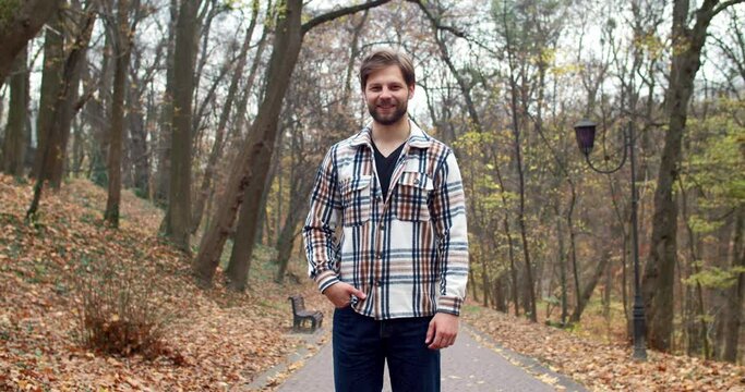 Portrait of successful stylish young man standing in park with trees leaves in autumn looking at camera smiling. Cute happy bearded handsome fashionable male in shirt and jeans on fresh air.