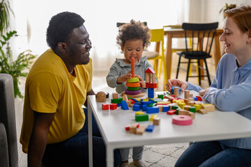 Mother and father supporting their cute little daughter in playing with colorful didactic wooden toys at home - obrazy, fototapety, plakaty