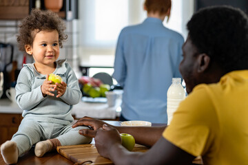 Multicultural family with a cute child at home kitchen. Parents feeding a small daughter with fruit...