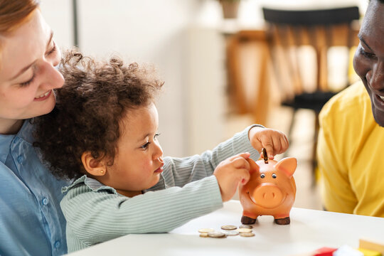 A Child Is Putting A Coin Into A Piggy- Bank. Savings Bank Accounts For Kids Concept. Child Saving Money For Future - Concepts. Multi-ethnic  Family