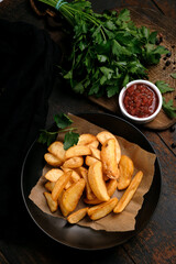 Fried potatoes in large pieces with ketchup in a black plate on a rustic wooden background, top view