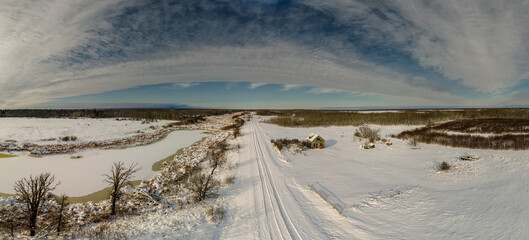 Aerial view of a snow covered marshy prairie scene with a narrow that has tire tracks in the snow.
