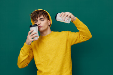  man stands on a green background in a yellow T-shirt and hat holding cardboard glasses for hot drinks in his hand and raises one to his mouth. Horizontal photo for inserting an advertising layout