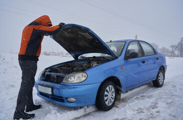 car broke down on the road in winter,a man opens the hood to repair a blue car in a blizzard