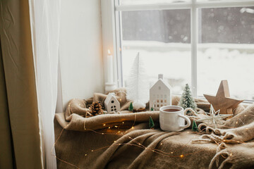 Winter hygge, Christmas still life. Warm cup of tea, christmas decorations, lights, little house,...