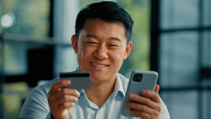 Happy Asian man client buyer businessman use banking mobile phone app at office hold plastic credit...
