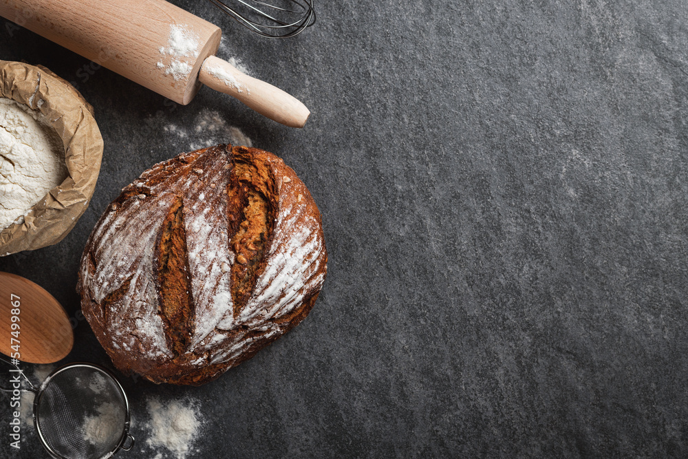 Poster whole loaf of fresh homemade bread with paper bag of flour on dark background with copy space top vi