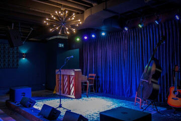 A music venue stage with stage lighting, microphone, amplifiers, PA system, upright piano, stand-up...