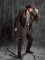 A young man in retro style, an adventure character. The hero of the adventure, a guy in a hat and a leather jacket, posing in the studio on a gray background - 547532097