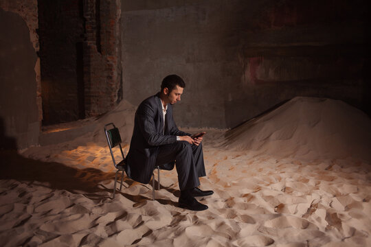 Economic crisis and recession, concept. A financier or businessman is sitting on a pile of sand in a room, Sad young man in a business suit lost everything, wasteland instead of business