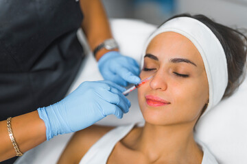 Fototapeta premium Portrait of an attractive caucasian European woman in her 30s wearing white protective hairband during collagen injection procedure done by a professional beautician in protective gloves. Aesthetic