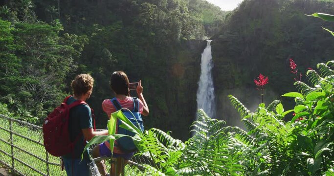 Travel vacation on Hawaii. Couple tourists on Hawaii by waterfall. Tourist taking photo using phone of Akaka Falls waterfall on Hawaii, Big Island, USA. Tourism concept with multicultural couple.