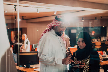 Portrait photo of Arab leader businessman with muslim hijab woman discussing business projects while using tablets and smartphone in modern glass office 