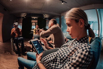 Pregnant woman sitting on the sofa of a modern office surrounded by colleagues and using a tablet